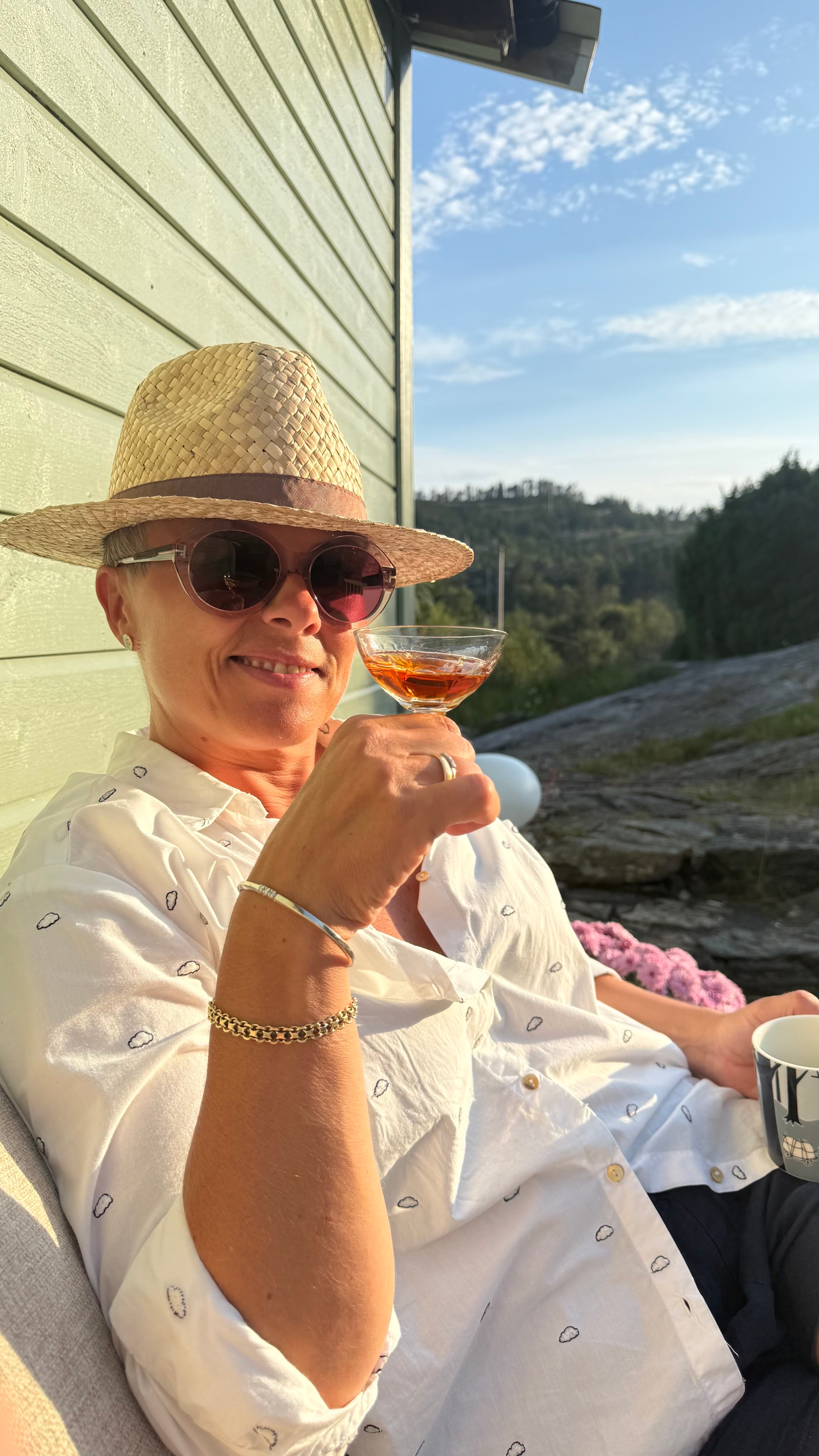 Johanne sitter ved hytteveggen i kveldssol med hatt og et glass i hånden.